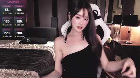 Media: Video of an East Asian woman with long black hair, wearing a black one-shoulder dress, sitting on a gaming chair in a dimly-lit bedroom.