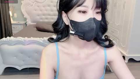 Video of a young Asian woman with fair skin, wearing a blue spaghetti-strap top, black mask, and shoulder-length black hair, sitting on a bed with a tufted white headboard in a light, elegant bedroom.