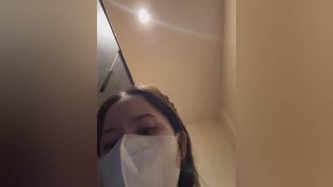 Media: A video of a young woman with long dark hair, wearing a white face mask, captured from a low angle in a narrow, dimly lit hallway. The background shows beige walls and a faint light source.