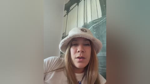 Video of a young woman with long, straight brown hair, wearing a white hat and white t-shirt, standing in front of a mural depicting a train track.