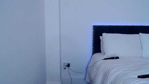 Video of a minimalist bedroom with a white bed, dark headboard, white pillows, and a nightstand with a black phone charger.