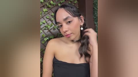 Media: A video of a young Asian woman with long, wavy black hair, wearing a black strapless dress and a choker necklace. She's standing against a wooden lattice covered in green ivy, with a blurred background.