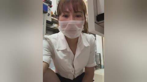 Media: A video of an East Asian woman with light skin and short, straight brown hair, wearing a white button-up shirt, a white mask, and dark pants, seated in a dimly lit room with shelves filled with medical equipment.
