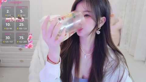 A video of a young Asian woman with long black hair, drinking from a plastic bottle in a bright, white bedroom with a digital display overlay.