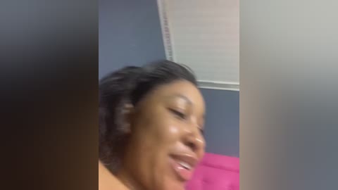 Video of a dark-skinned Black woman with short, curly hair, laughing with eyes closed, in a room with blue walls and a pink bedspread.