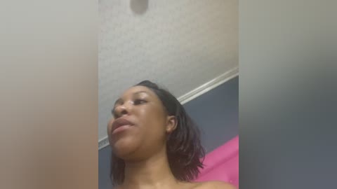 Video of a young Black woman with medium-brown skin, shoulder-length curly hair, and full lips, standing indoors, wearing a pink top, with a textured white ceiling and blue wall in the background.