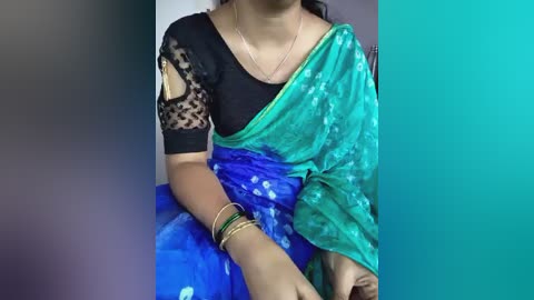 Video of a woman in traditional Indian attire: black lace blouse, turquoise saree, green bangles, sitting indoors.