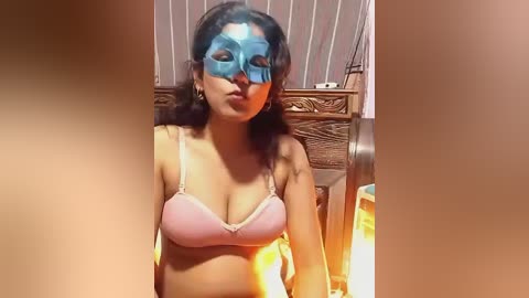 Video of a woman with medium skin tone, wearing a blue superhero mask and pink bra, standing in a dimly lit room with a wooden headboard.