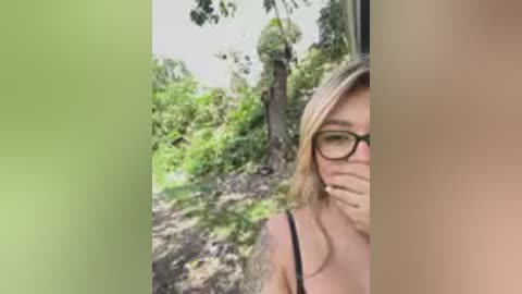 Video of a blonde woman with glasses, partially obscured by a green vertical gradient, standing in a lush, green forest. She has a thin build and is wearing a black tank top.