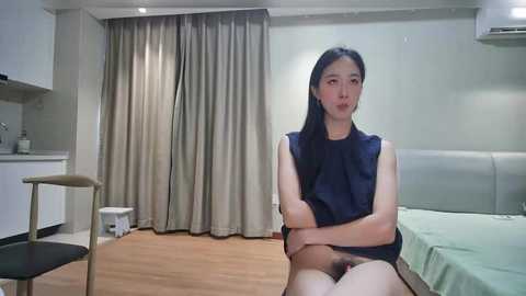 Video of a young East Asian woman with long black hair, wearing a sleeveless navy top, sitting on a bed in a modern, minimalist bedroom with beige curtains and wooden floor.