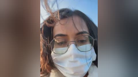 Video of a young woman with medium brown skin, wearing glasses and a white surgical mask, her dark hair slightly tousled, against a blurred, outdoor background.