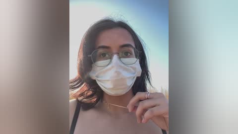 Video of a woman with medium-length dark hair, wearing round glasses, a white surgical mask, and a black top, standing outdoors against a blurred blue sky background.