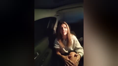 Media: A video shows a woman with long hair, wearing a white sweater, sitting inside a dark, shadowy car, holding a brown bag in her lap.