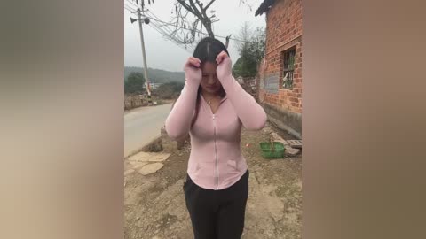 Media: Video of a woman in a pink hoodie and black pants adjusting her black hat outdoors on a rural dirt road, surrounded by brick buildings and bare trees.