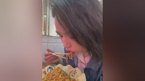Media: A video of a woman with long dark hair, wearing a denim jacket, eating noodles with chopsticks in a tiled kitchen.
