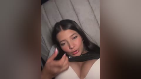 Video of a young woman with long black hair, fair skin, wearing a black bra and white top, talking on a black phone, against a grey fabric backdrop.