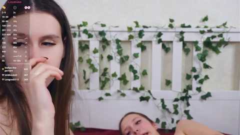 Video of a young woman with long brown hair and fair skin, holding her finger to her lips, in a bedroom with a white crib adorned with green ivy.