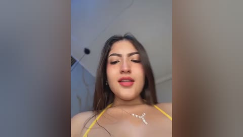 A video of a young woman with long dark hair, wearing a yellow bikini top, standing indoors. She has a light skin tone, full lips, and is smiling.