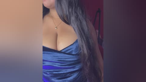 Video of a woman with long, straight black hair wearing a strapless, deep blue satin dress that accentuates her large breasts. She has a light brown complexion and wears a delicate gold necklace. Background is a gradient of reds and purples.