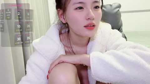 A video of a young East Asian woman with fair skin, dark hair, and red lipstick, wearing a white plush robe, sitting on a black leather chair in a modern indoor setting.