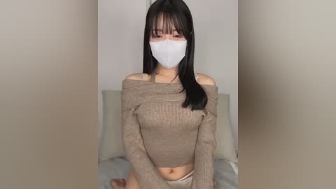 Media: A video of an East Asian woman with long black hair, wearing a beige off-shoulder sweater, white mask, and beige underwear, seated on a bed in a simple, light-colored room.