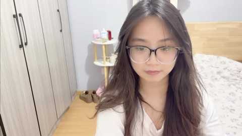 Video of an Asian woman with long black hair, wearing glasses, white top, standing in a bedroom with light wooden furniture, toiletries on a white shelf, and a bed with white sheets.