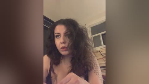 A video of a young woman with curly, voluminous hair, wearing a black tank top, looking surprised with her mouth open, standing in a room with beige walls and a bookshelf in the background.