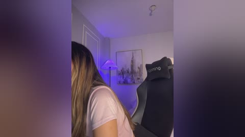 A video of a woman with long, straight, blonde hair wearing a white T-shirt, seated in a black gaming chair, in a dimly lit room with a lit lamp and cityscape artwork on the wall.