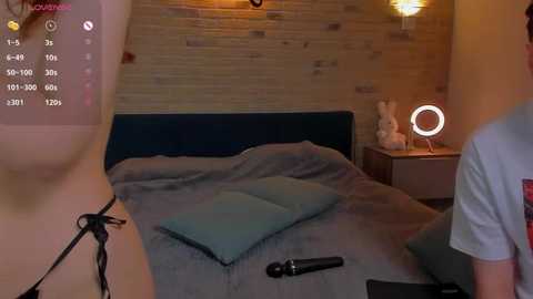 Video of a modern, cozy bedroom with a plush bed, teal pillows, wooden wall, and a circular mirror light on a nightstand. A man's torso and a black microphone are visible.