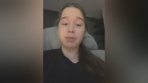 Video of a young woman with fair skin and long brown hair, wearing a black top, looking straight at the camera with a neutral expression. Background features a white pillow and dark furniture.