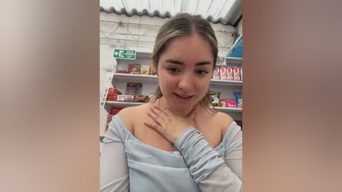 Video of a young Caucasian woman with light skin and brown hair, wearing a light blue off-the-shoulder top, touching her chest in a grocery store with shelves of products in the background.