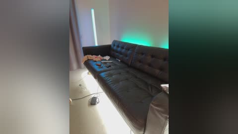 Video of a dimly lit, minimalist room with a black leather sofa against a beige wall. Neon green light casts a soft glow on the sofa. Clothes and shoes are scattered on the floor, suggesting a lived-in, casual atmosphere.