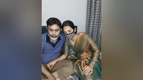 Media: Video of a man in a blue shirt and beige pants with a bandana, sitting on a sofa next to a woman in a green sari and mask, both appearing distressed in a dimly lit room.
