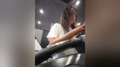 Media: Video of a woman with long, dark hair wearing a white t-shirt, driving a black car in a dimly lit, modern interior.