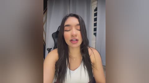 Video of a young woman with long black hair, wearing a white tank top, standing with her eyes closed, smiling, indoors near a window with gray curtains.
