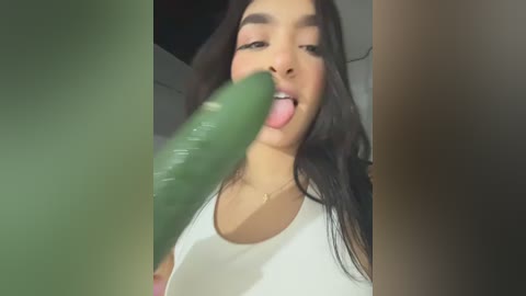 Video of a young woman with long dark hair, light brown skin, and a white tank top, licking a large, green dildo. The background is out of focus.