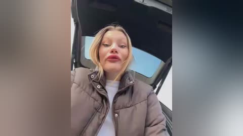 Media: Video of a blonde woman with light skin, wearing a brown puffer jacket and white shirt, sitting in a car with the door open, looking forward with a neutral expression.