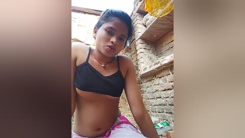 Video of a young woman with light brown skin, wearing a black sports bra and pink shorts, sitting on a dirt floor between brick walls in a rural setting.