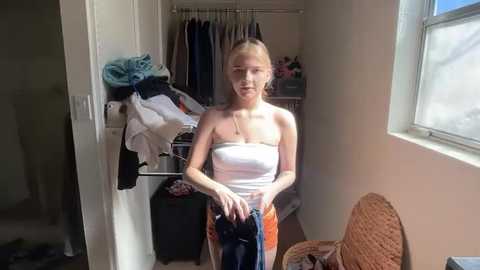 Media: Video of a young blonde woman in a white strapless top and orange shorts, holding a pair of jeans, in a cluttered bedroom with a wicker chair, clothes on racks, and sunlight streaming in.