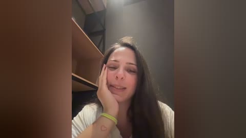 Video of a young woman with long brown hair, wearing a white top, resting her head on a wooden surface with a green bracelet.