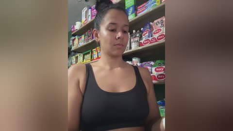 Video of a young woman with dark hair in a bun, wearing a black sports bra, standing in a colorful, cluttered supermarket aisle filled with various packaged products.