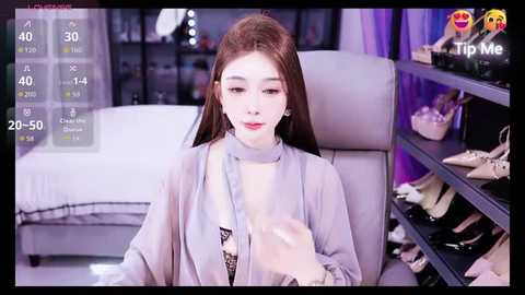 Video of a young Asian woman with long brown hair, fair skin, and a slender figure, wearing a light grey robe, sitting in a modern living room with shelves filled with shoes.
