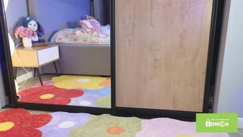 Video of a bedroom with a wooden wardrobe door reflecting a bed covered with colorful pillows and a flower-patterned rug. The room has a playful, child-friendly atmosphere.