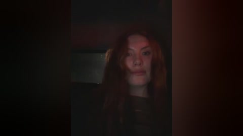 Video of a young woman with long, curly red hair peering through a dark doorway, wearing a dark outfit. The background is dimly lit, creating a mysterious, suspenseful atmosphere.
