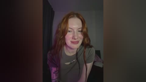 A video of a smiling, fair-skinned woman with long, wavy red hair, wearing a gray shirt, standing in a dimly lit room with dark curtains.