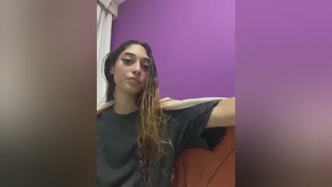 A video of a young woman with long, wet, dark brown hair, wearing a dark t-shirt, leaning against a purple wall and orange couch.