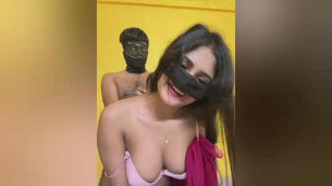 Video of a smiling woman with long black hair and large breasts, wearing a pink bra and black face mask, standing in front of a man in a black mask and a yellow background.
