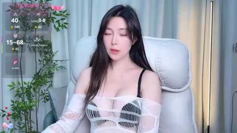 Media: A video of an East Asian woman with long black hair, wearing a sheer white lace dress, sitting on a white leather chair. The background features a lush green plant and a digital temperature display.