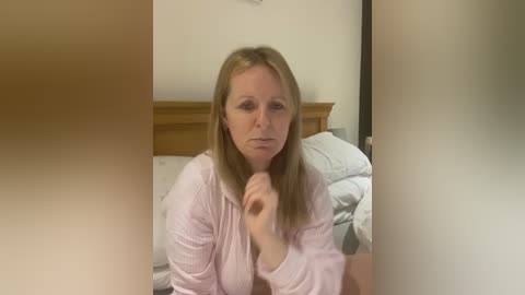 Media: Video of a middle-aged Caucasian woman with long blonde hair, wearing a light pink robe, sitting on a bed with white bedding and a wooden headboard. She looks contemplative, hands clasped.