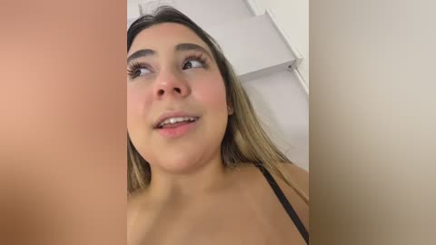 Video of a young woman with light brown skin, shoulder-length brown hair, and large eyes. She has a slight smile and is wearing a black strapless top. The background is a white wall with a door handle.